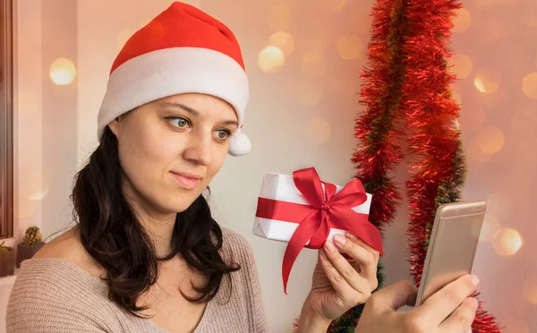A girl in a Santa hat holds a gift box and shows it to a smartphone. Tinted photo with bokeh added. Merry christmas and happy new year greetings online