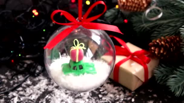 Oyuncak arabalı şeffaf bir Noel ağacı oyuncağı ve arka planda yağan karın altında bir Noel ağacı ve bir Noel ağacı.. — Stok video