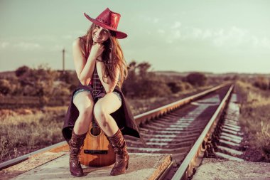 genç gülümseyen brunet gitarda yaz demiryolu günbatımında oturan şapkalı