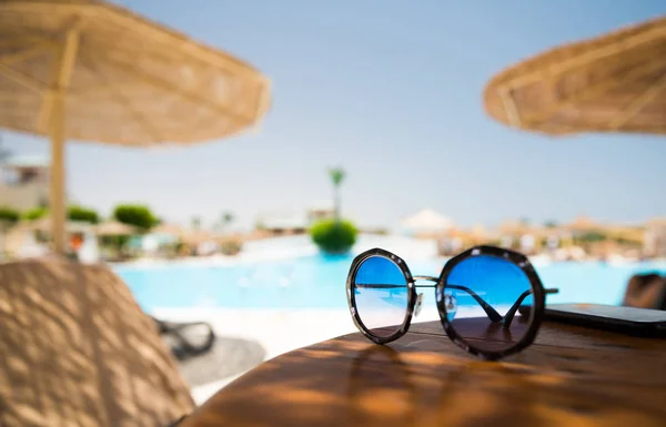 sunglasses on summer wooden table with pool at background