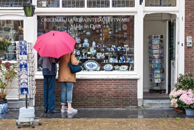 Delft, Hollanda - 25 Ağustos 2018: Hollanda Delft, Hollanda 'da el boyaması geleneksel çömlek dükkanının vitrinine bakan kırmızı şemsiyeli iki turist