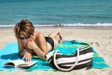 Orta yaşlı, beyaz, bikinili bir kadın kumsalda uzanmış, parlak bir yaz gününde kitap okuyor. 