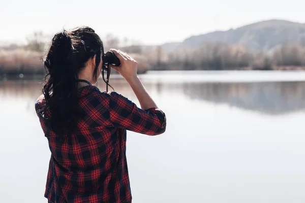 带着双筒望远镜和格子衬衫的湖上码头上的年轻黑发女子 — 图库照片