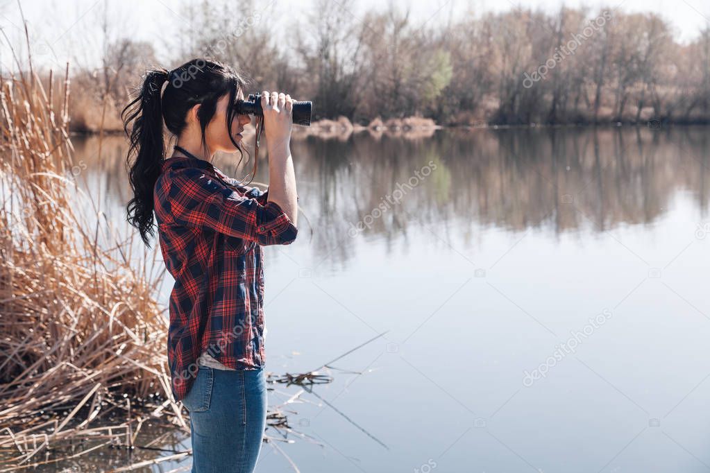 Young brunette woman on a lake's jetty with binoculars and plaid shirt