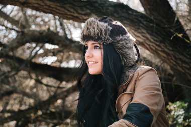 Young attractive woman in leather bomber hat smiling and looking away in forest   clipart