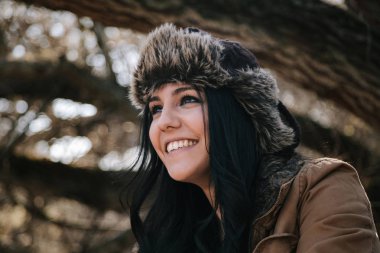 Young attractive woman in leather bomber hat smiling and looking away in forest  clipart