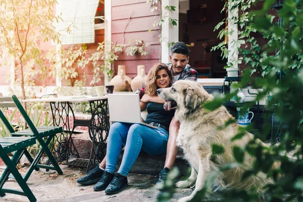Genç çift ve bahçe sandalye ahşap evlerinin bir dizüstü bilgisayar ile gülümseyen onların köpek oturma