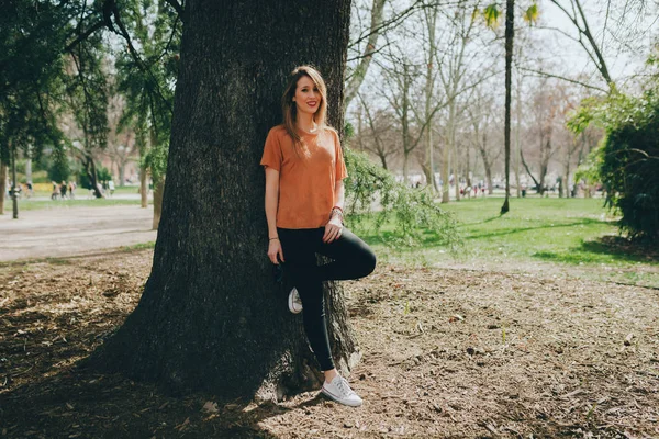 Jeune femme blonde souriante soutenue sur un arbre avec un jean noir — Photo