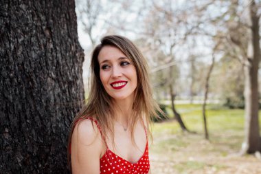 Kırmızı elbiseli bir ağaç karşı eğilim genç gülümseyen sarışın kadın