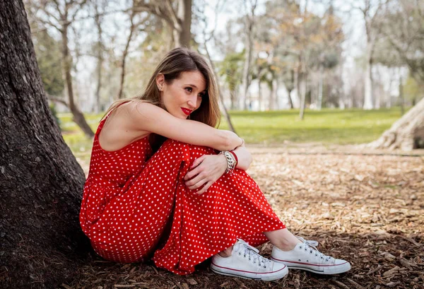 Giovane sorriso donna bionda seduta vicino all'albero con abito lungo rosso — Foto Stock