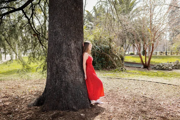 Mladá usměvavá blondýnka s červených šatech opřený o strom — Stock fotografie