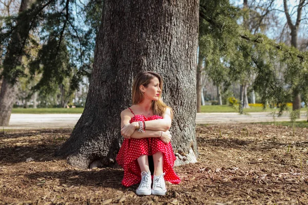 Joven mujer rubia seria sentada cerca del árbol con vestido largo rojo — Foto de Stock