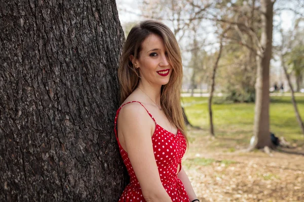 Junge blonde Frau mit rotem Kleid lehnt an einem Baum — Stockfoto