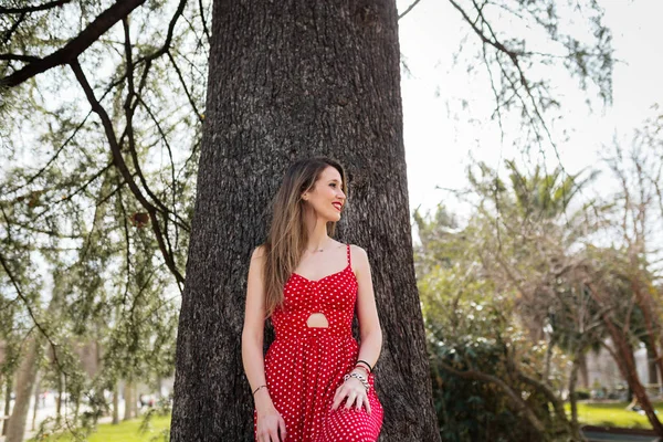 Jeune femme blonde souriante avec robe rouge appuyée contre un arbre — Photo