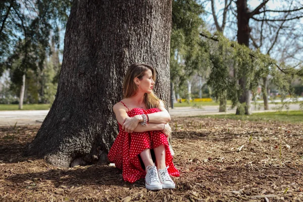 Junge ernsthafte blonde Frau sitzt in der Nähe von Baum mit rotem langen Kleid — Stockfoto