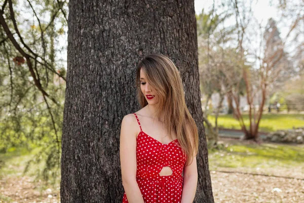 Jeune femme blonde souriante avec robe rouge appuyée contre un arbre — Photo