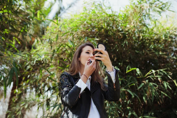 Young blonde woman wearing a leather jacket makes up in the park — Stock Photo, Image