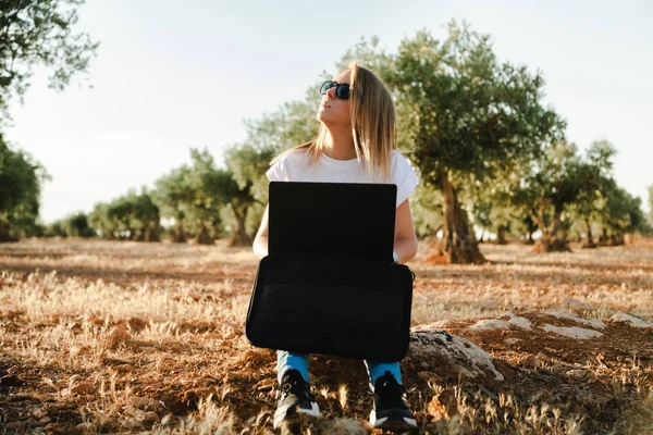 Młoda kobieta z futerał na laptopa siedzi w polu drzewa oliwnego — Zdjęcie stockowe
