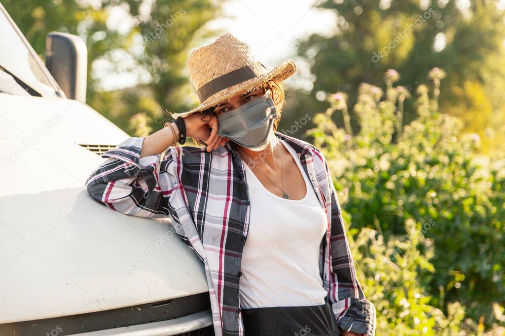 Young farmer woman with straw hat and surgical mask leaning against the van and holding her hat