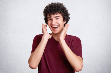 Neşeli kıvırcık genç erkek iyi ruh hali, gülüyor mutlu olmak hoş şaka, gülümsüyor, duyar gibi pozlar beyaz beton duvara rahat t gömlek giyer. İnsanlar, mutluluk ve positiveness kavramı