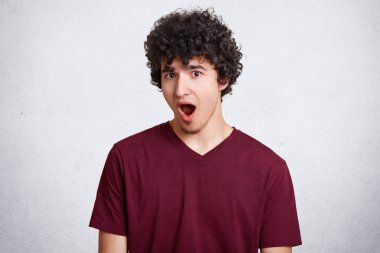 Hayret çekici kıvırcık genç erkek görünümü ile korkmuş ifade, tutar duymak hayrete ağız geniş açıldı, Haberler, şok edici beyaz beton duvara teşkil etmektedir. İnsanlar, surprisement kavramı