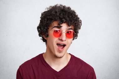 Headshot hayran kıvırcık erkek moda güneş gözlüğü, rahat kıyafetler giyer, beklenmeyen News, pozlar beyaz beton duvara hayrete açtı, ağız tutar. İnsanlar, moda ve gençlik kavramı