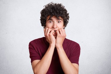 Korkmuş nevrotik erkek ısırıkları parmak çivi, kameraya bakıyor, gevrek saçlı, rahat kıyafetler, pozlar beyaz beton duvara giyer. Korkmuş genç kıvırcık saçlı panik içinde olmak. Duygular kavramı