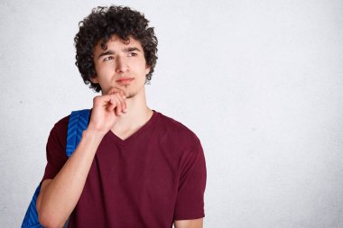 Kıvırcık düşünceli erkek çene tutar, sırt çantası taşıyan, düşünceli mesafe içine görünüyor, boşluk ile beyaz arka plan üzerinde izole Üniversitesi'nde derslere bulunmadan önce yaz tatili hakkında düşünüyor