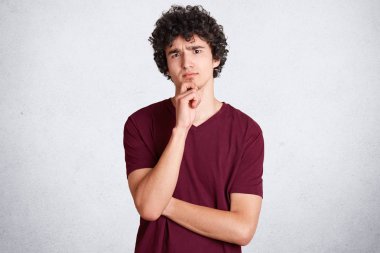 NET saçlı yakışıklı düşünceli erkek model portresi çene tutar ve ifade sıktı, rahat t gömlek giyer, beyaz beton duvara karşı duruyor. Genç hakkında bir şey düşünen