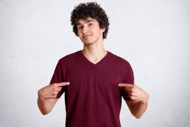 Attractive male youngster with crisp dark hair, points at casual t shirt, shows free space for your logo or advertisement, isolated over white concrete wall. People, clothing, design concept clipart