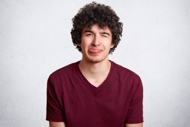 Neşeli mutlu genç kıvırcık erkek gözleri yanıp söner ve gösterir dil, kameraya, yüz buruşturma yapar vardır komik ifade, beyaz arka plan üzerinde izole rahat kıyafetler giymiş. Genç kapalı eğlenir