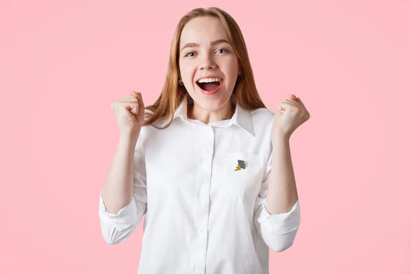 Successful beautiful female student clenches fists, celebrates and rejoices well done project work or final exam, being in high spirit, ready to have holidays, wears formal white shirt, isolated