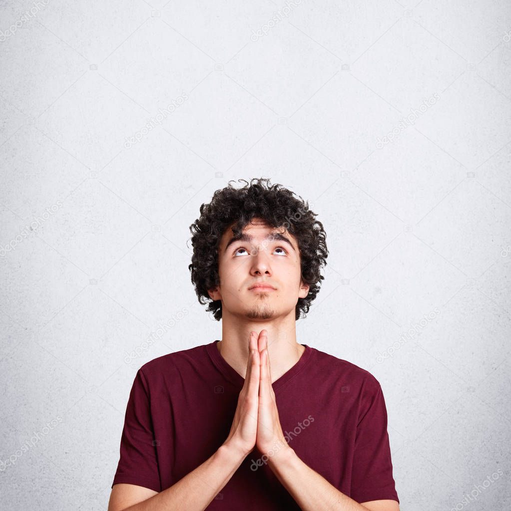 Vertical shot of pleasant looking miserable male prays for something, looks upwards with great hope, asks for health, poses against white background with blank space for your advertising content