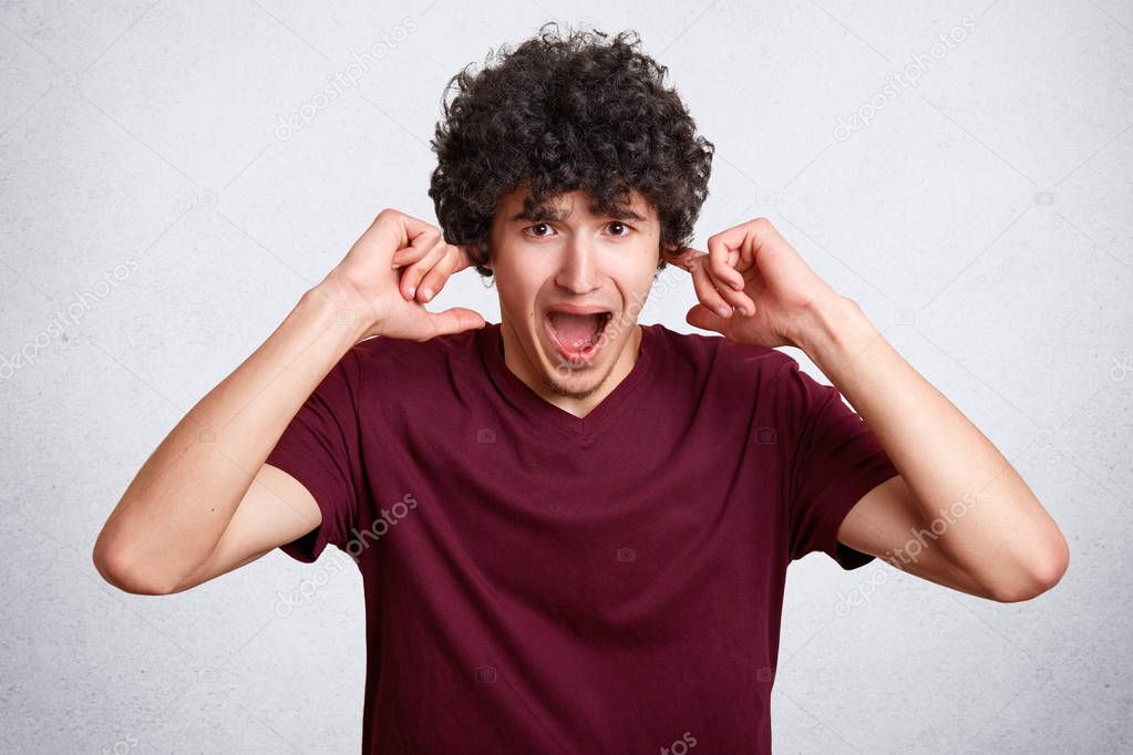 Stressful curly male plugs ears, being in panic, avoids loud irritating sound, wears casual t shirt, stands against white background. Annoyed handsome teenager doesn`t want to hear pop music.
