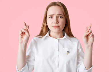 Güzel güzel beyaz kadın parmak çarpı işareti tutar, büyük umutla kameraya benziyor, pembe bir arka plan üzerinde izole casual beyaz t gömlek giyer. Güzel öğrenci final sınavı geçirmeden önce dua ediyor