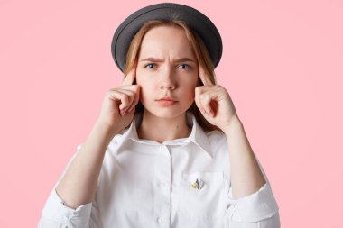 Headshot düşünceli ifade ile ciddi kadın parmaklarını tapınak üzerine tutar, trendy şapka ve zarif beyaz gömlek giyer, pembe arka plan izole kafasında bir şey hatırlamak çalışır
