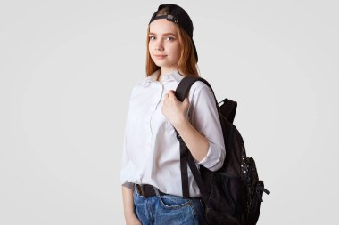 Şık siyah şapkalı ciddi sevimli genç kapalı atış sırt çantası taşıyan, beyaz gömlek ve beyaz arka plan üzerinde izole kot giyer. Güzel kız öğrenci okula hazırlar. Eğitim kavramı