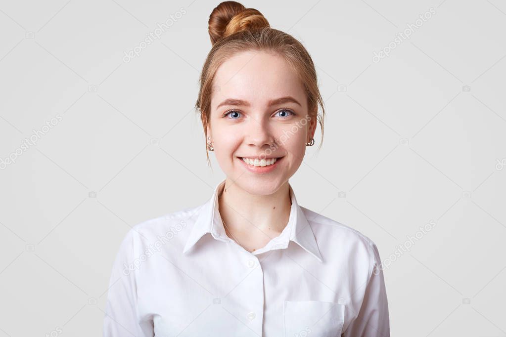 Indoor shot of beautiful female with pleasant smile, hair knot, being in good mood, wears stylish formal white shirt, isolated over studio background. People, happiness, positiveness concept