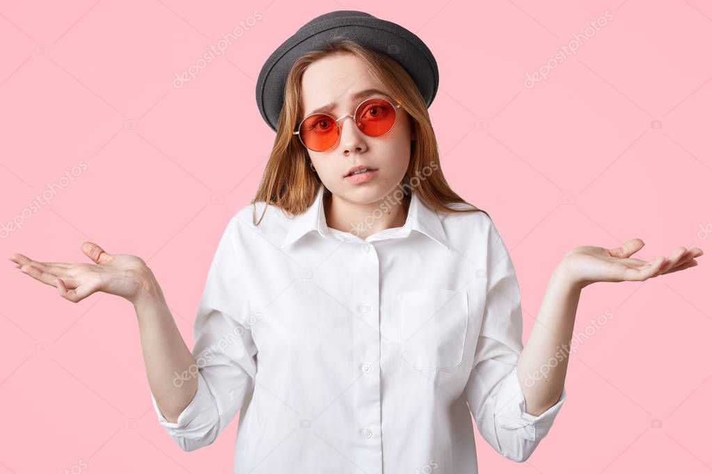 Hesitant beautiful hipster female shrugs shoulders in bewilderment, being uncertain and has clueless expression, wears stylish hat and red sunglasses, poses against pink background. Who cares?