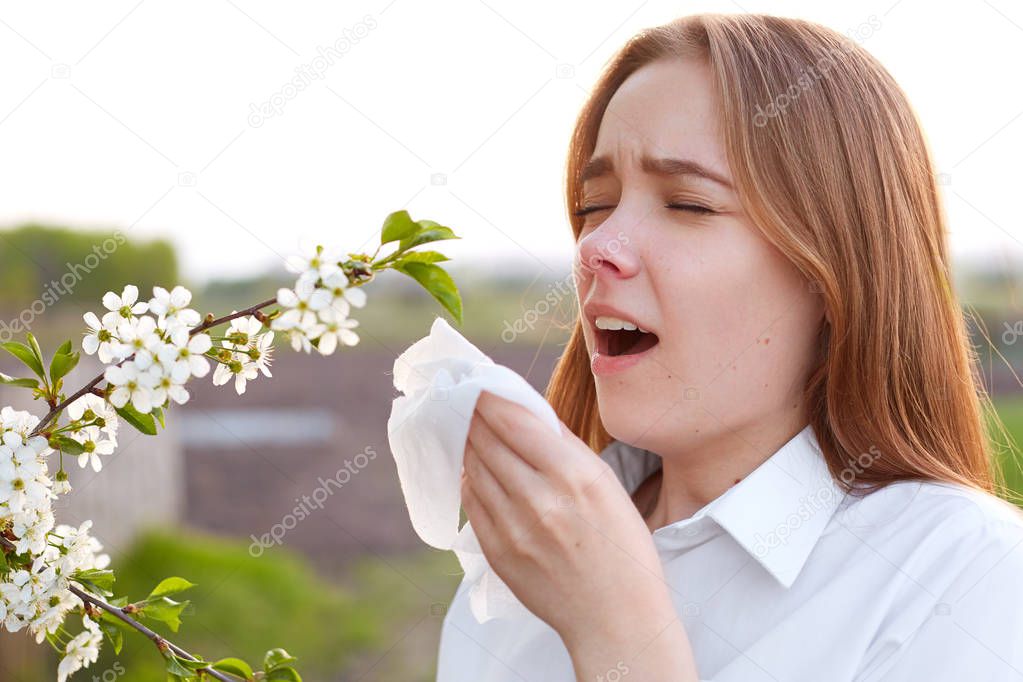 Seasonal allergy. Pretty young female blows nose and sneezes, stands in front of blooming tree, being allergic to blossom, holds handkerchief. Lovely woman has allergy during spring near allergens