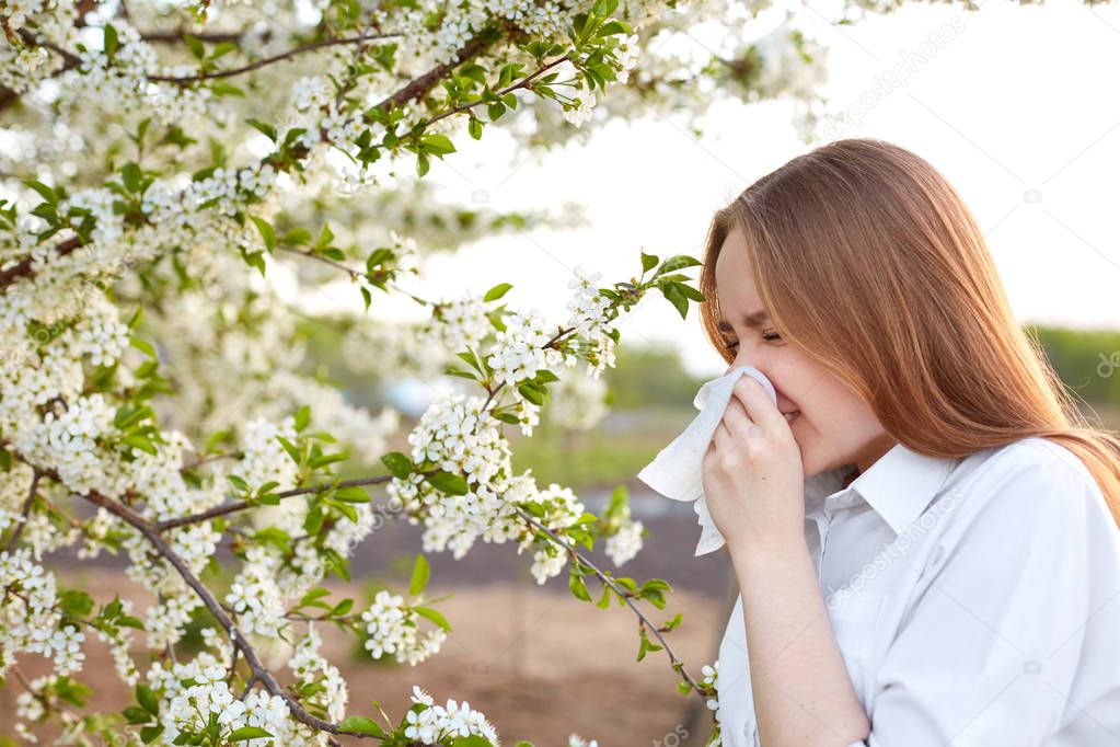 Pollen allergy symptom. Sideways outdoor shot of young European female sneezes in handkerchief or blows in wipe , being allergic to blossom during spring, stands on front of blooming tree outside.