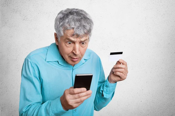 惊讶的老男人用灰白的头发盯着智能手机 不懂什么东西 拿着手机和信用卡 隔着白色的混凝土背景 巨大的困惑和 Omg 的概念 — 图库照片