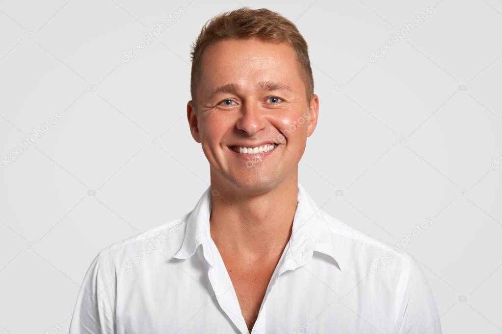 Cheerful satisfied male with positive expression, shows white teeth, being in good mood after successful meeting, dressed in white shirt in one tone with background. People and lifestyle concept