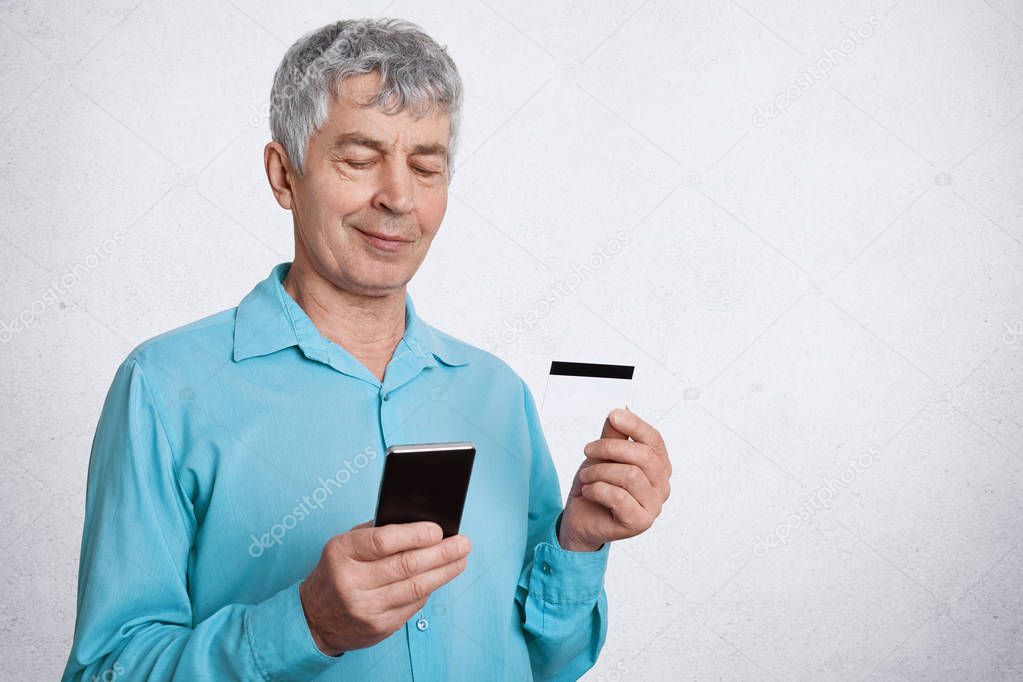 Elegant male pensioner holds smart phone and credit card, checks his account and pension in online webpage, satisfied to recieve much money poses against white concrete wall. Elderly man does shopping