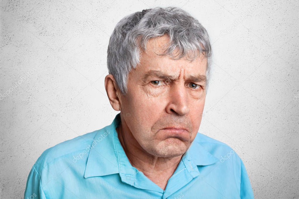 Displesed unhappy wrinkled mature male frowns face and curves lips, wears formal shirt, poses against white concrete background. Close up of elederly man dissatisfied with ammount of pension