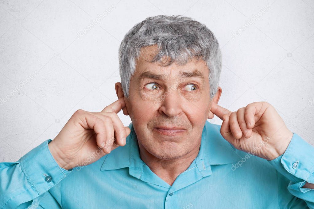 Headshot of wrinkled elederly male with grey hair plugs ears as doesn`t want hear something, looks with unhappy expression away, ignores someone, isolated on white concrete wall. I don`t want hear it