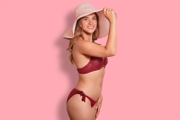 スリムな美しいフィットのボディを持つ女性 スタンド横にカメラに笑顔夏帽子と水着を着て ピンクの背景に分離 夏を楽しんでいます ライフ スタイル 休暇の概念 — ストック写真