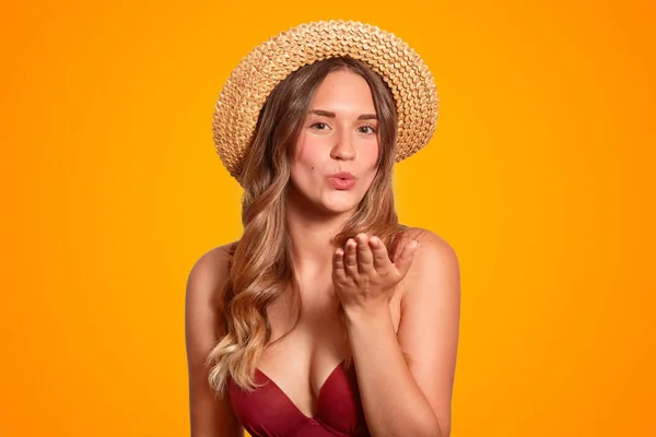 Horizontal shot of pleasant looking female with healthy tanned skin, wears summer hat and swimsuit, makes air kiss gesture, flirts with handsome guy at seaside, isolated over bright studio background