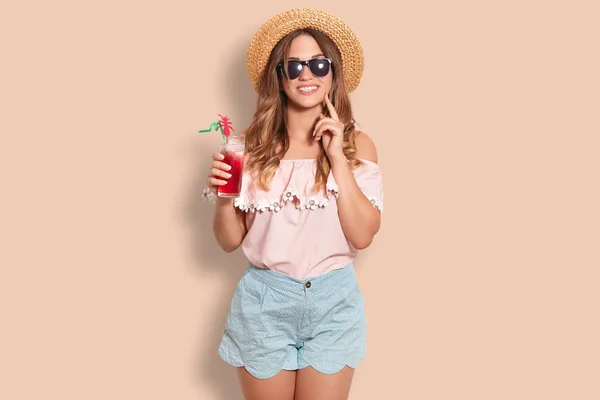 正面女性在夏天帽子 窗帘上衣和短裤 举行冷的新鲜的鸡尾酒 去在海滩 有友好的微笑 站立反对米色背景 有乐趣 人和夏天时间概念 — 图库照片