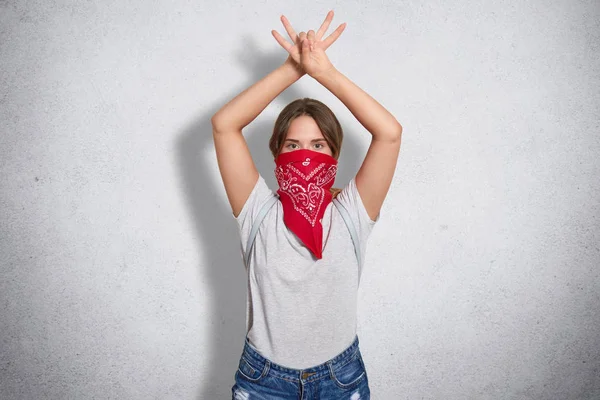 Young cute female feminist wears red bandana and casual grey t shirt, protects womens rights, keeps hands raised and crossed, looks seriously, stands against concrete wall. Feminism concept.